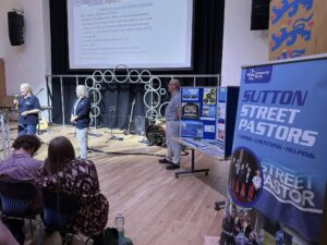 Sutton Community Works staff and volunteer on stage at a church visit.